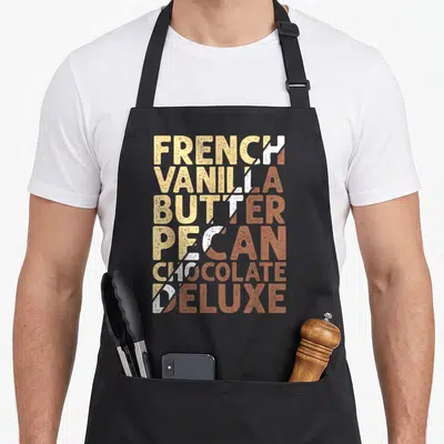 French butter apron - Stacked text FRENCH VANILLA BUTTER PECAN CHOCOLATE DELUXE split diagonally in beige white — shown on a black cotton canvas apron against a white background