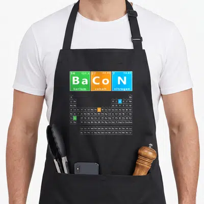 Bacon table apron - Periodic table BaCoN pun with green barium, orange cobalt, blue nitrogen tiles above grey chart — shown on a black cotton canvas apron against a white background