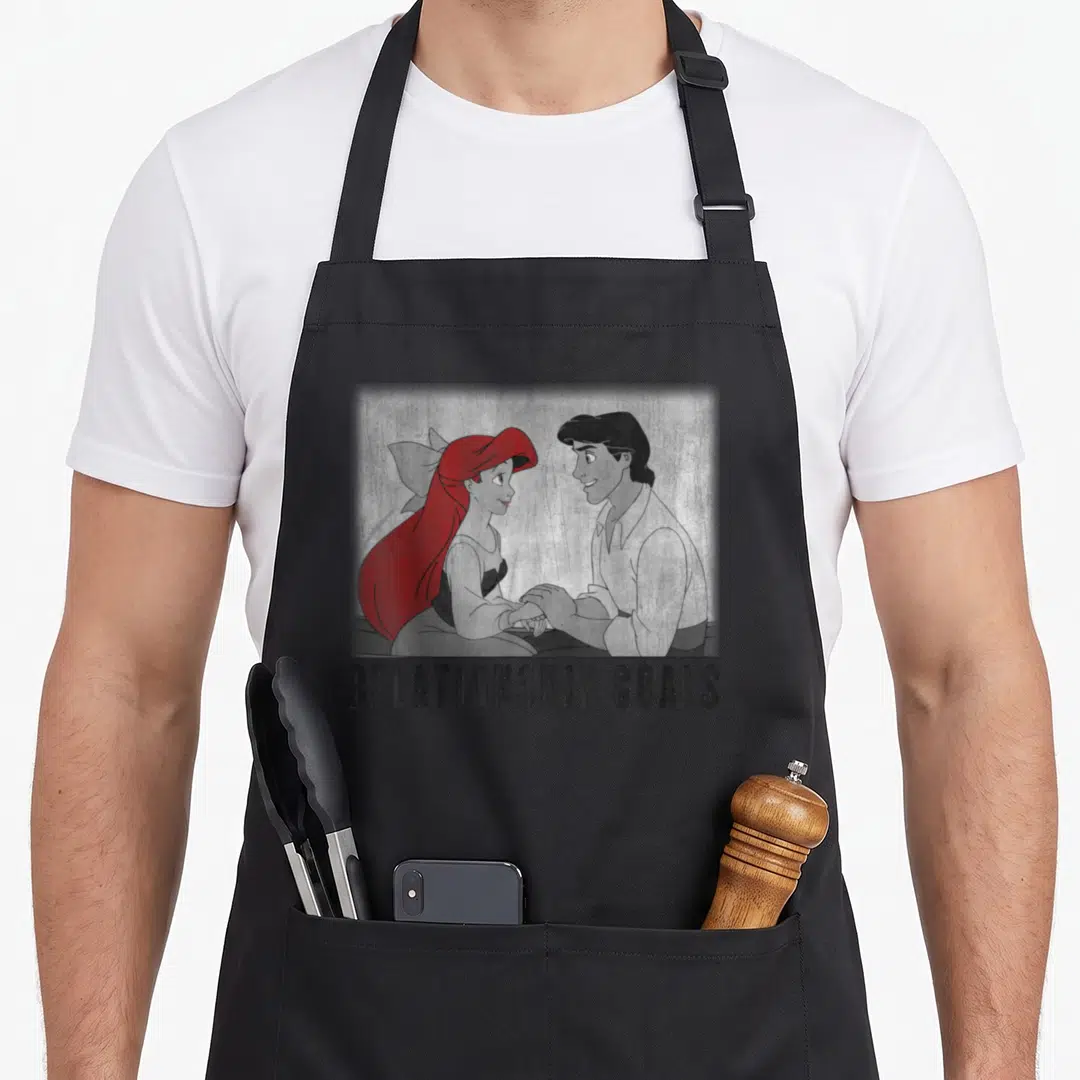 Relationship mermaid apron - Cartoon couple holding hands with red hair and RELATIONSHIP GOALS text below — shown on a black cotton canvas apron against a white background