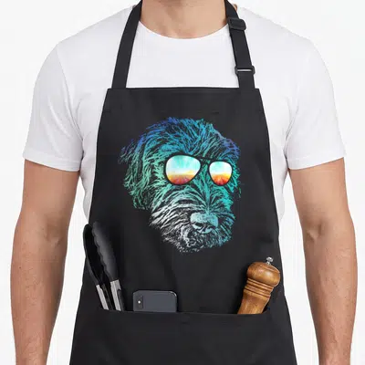 Wirehaired dog apron - Stylized wirehaired dog head in blue teal orange and white wearing sunglasses reflecting a sunset — shown on a black cotton canvas apron against a white background