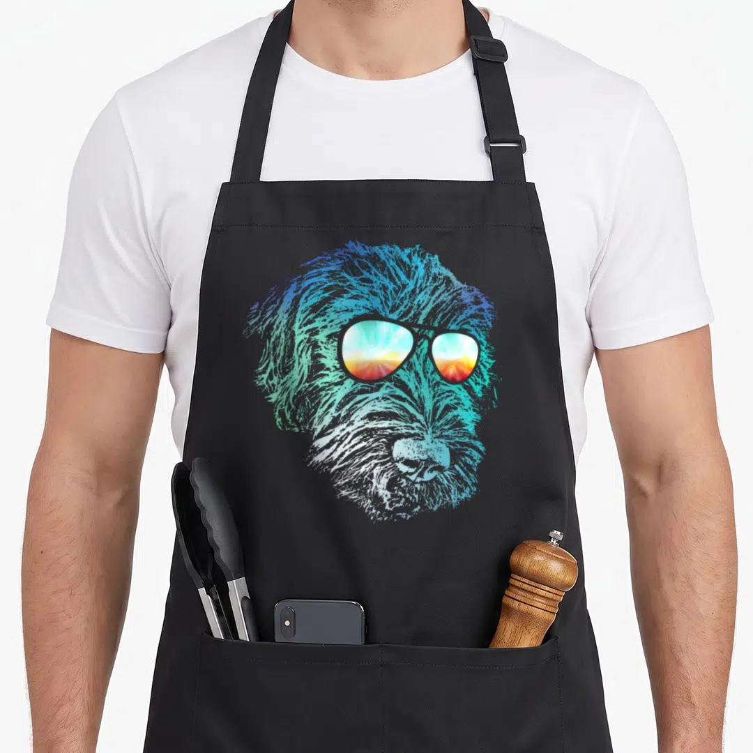 Wirehaired dog apron - Stylized wirehaired dog head in blue teal orange and white wearing sunglasses reflecting a sunset — shown on a black cotton canvas apron against a white background