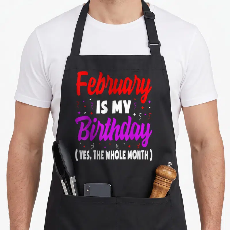 February whole month birthday apron - Red purple and white text reading February is my birthday yes the whole month — shown on a black cotton canvas apron against a white background