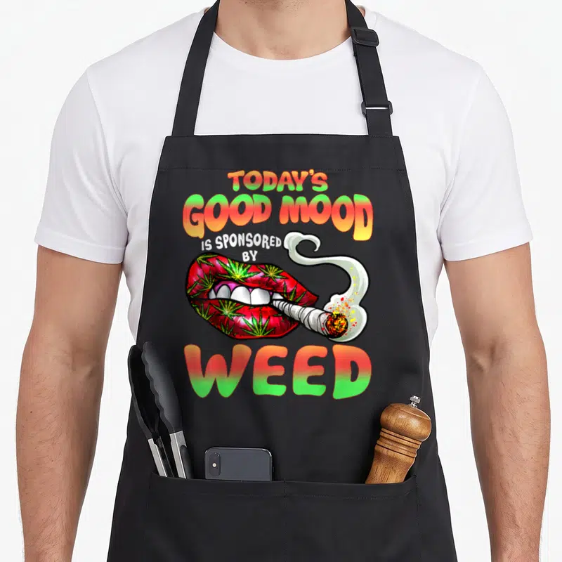 Lip smoking apron - TODAY'S GOOD MOOD IS SPONSORED BY WEED text above red lips with green leaves and a smoking joint — shown on a black cotton canvas apron against a white background