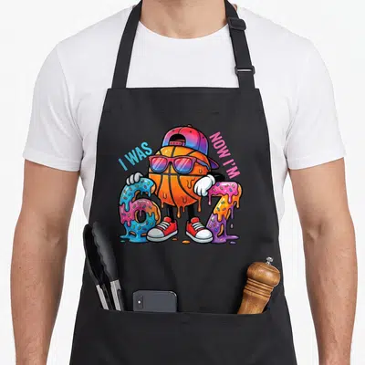 Birthday basketball meme apron - Cartoon basketball in cap between melting 6 and 7 numbers with I WAS NOW I'M text — shown on a black cotton canvas apron against a white background