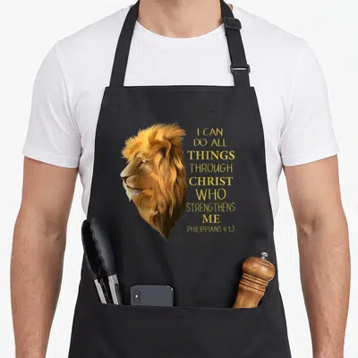 Lion christian apron - Realistic lion head in profile next to stacked gold text of Philippians 4:13 verse — shown on a black cotton canvas apron against a white background