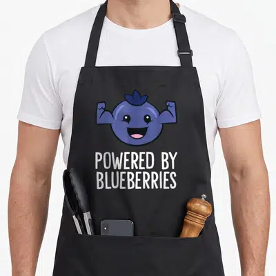 Powered blueberries apron - Cartoon blueberry flexing arms above POWERED BY BLUEBERRIES text in white — shown on a black cotton canvas apron against a white background
