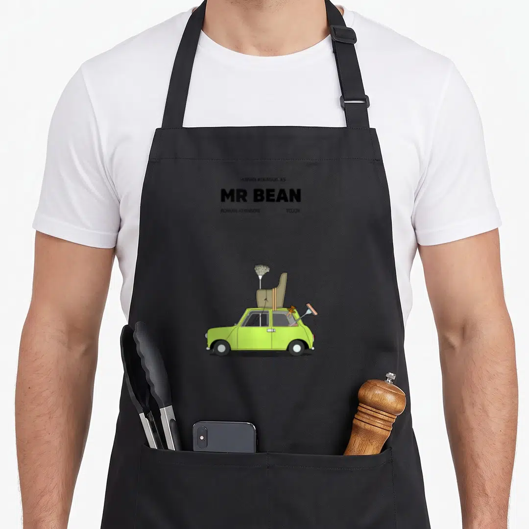 Movie mr apron - Lime green car with armchair mop and Teddy on roof under movie poster text — shown on a black cotton canvas apron against a white background