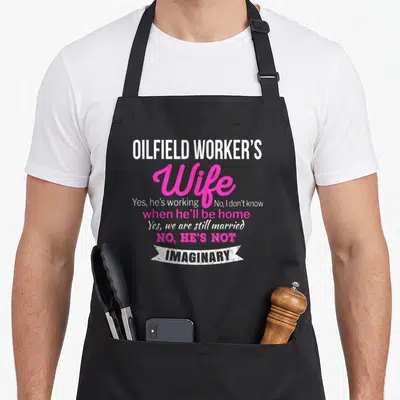 Wedding wife apron - White and pink text reading OILFIELD WORKER'S Wife Yes we are still married NO HE'S NOT IMAGINARY — shown on a black cotton canvas apron against a white background