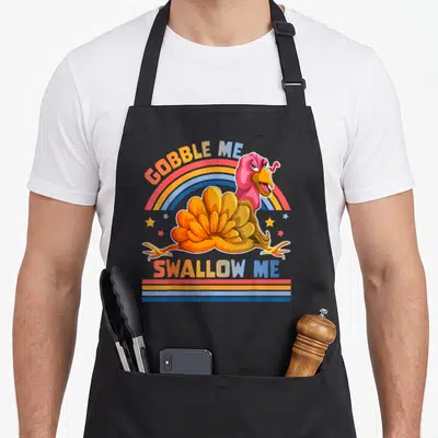Gobble swallow thanksgiving apron - Cartoon turkey with rainbow arch and GOBBLE ME SWALLOW ME text in orange pink blue — shown on a black cotton canvas apron against a white background