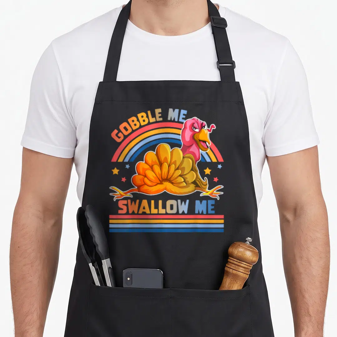 Gobble swallow thanksgiving apron - Cartoon turkey with rainbow arch and GOBBLE ME SWALLOW ME text in orange pink blue — shown on a black cotton canvas apron against a white background