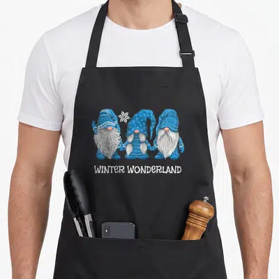Wonderland winter apron - Three blue gnomes with white snowflake and WINTER WONDERLAND text below — shown on a black cotton canvas apron against a white background