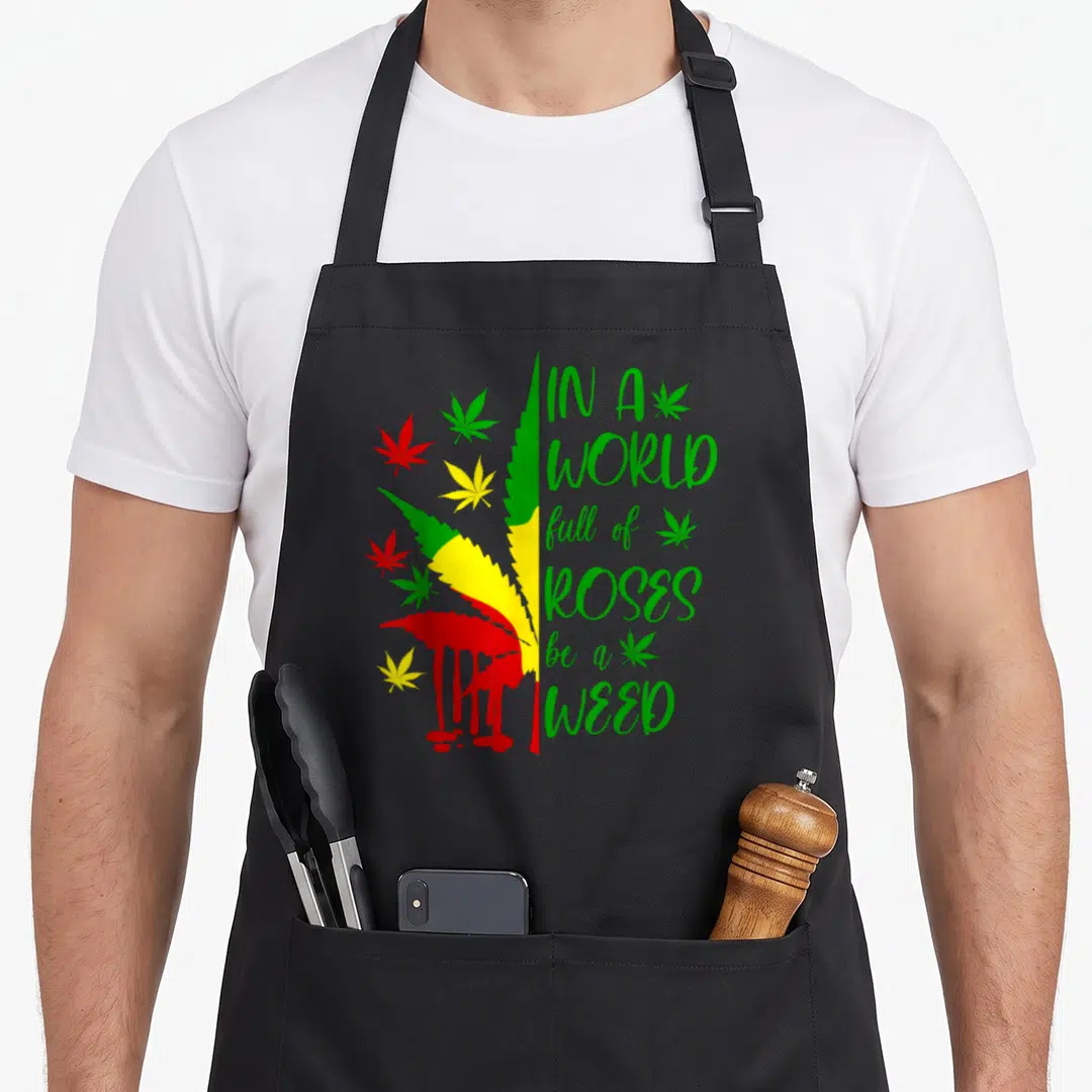 Weed rose apron - Tri-color cannabis leaf with dripping red paint and stacked green text reading IN A WORLD full — shown on a black cotton canvas apron against a white background