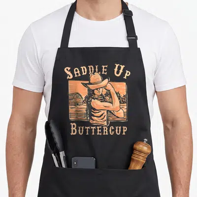 Saddle western aprons - Retro cowgirl with crossed arms in peach frame with Saddle Up Buttercup text — shown on a black cotton canvas apron against a white background