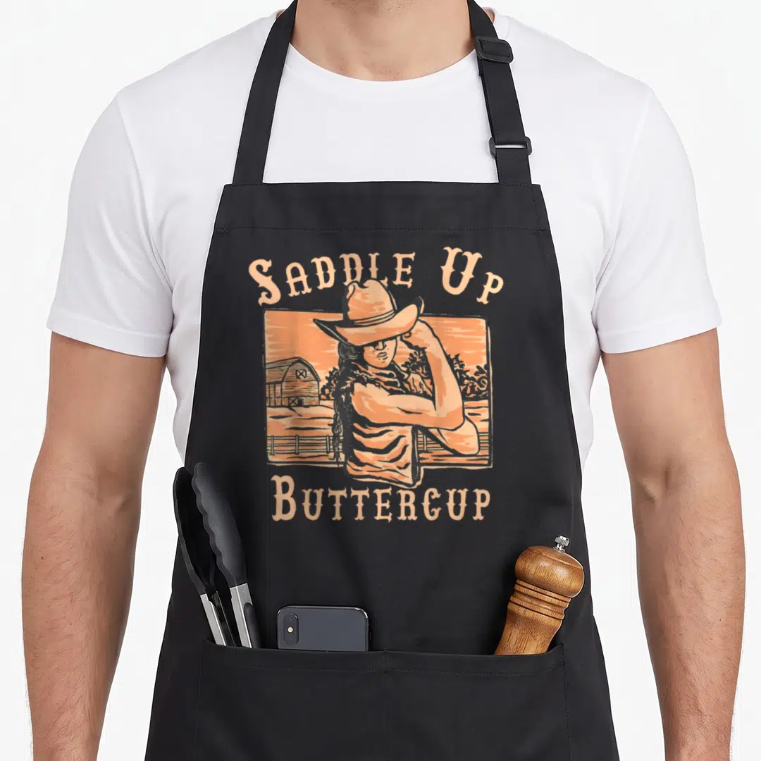 Saddle western aprons - Retro cowgirl with crossed arms in peach frame with Saddle Up Buttercup text — shown on a black cotton canvas apron against a white background