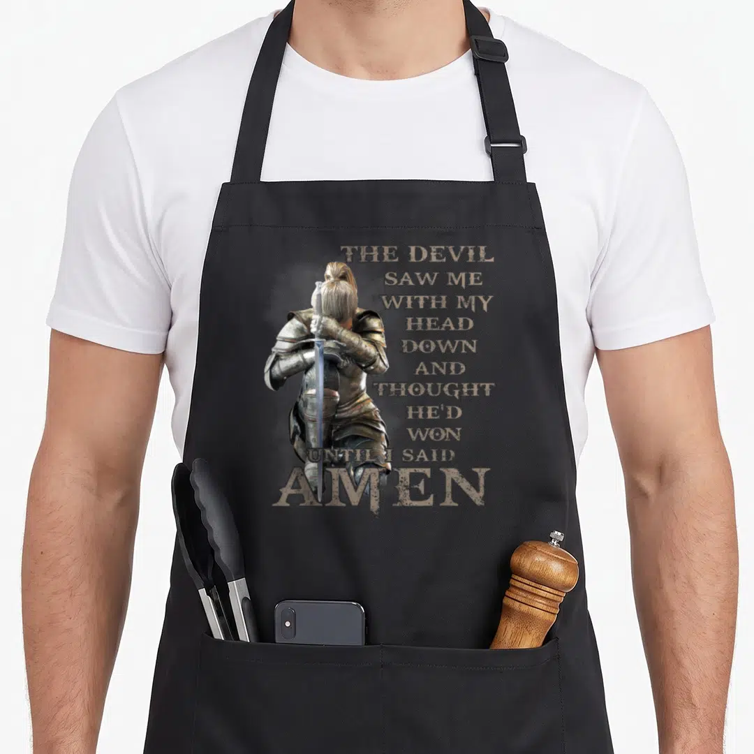 Amen devil's apron - Kneeling knight in silver armor holding sword with vertical tan text reading THE DEVIL SAW ME — shown on a black cotton canvas apron against a white background