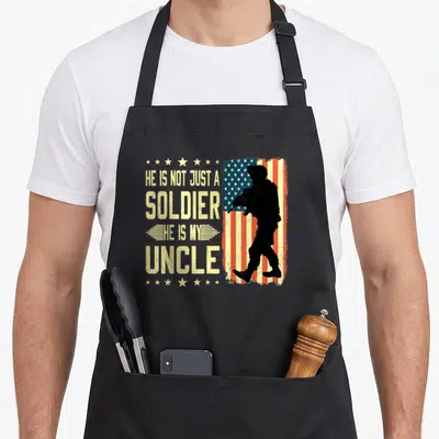 Uncle military apron - Soldier silhouette on distressed flag with He Is Not Just A Soldier He Is My Uncle text — shown on a black cotton canvas apron against a white background