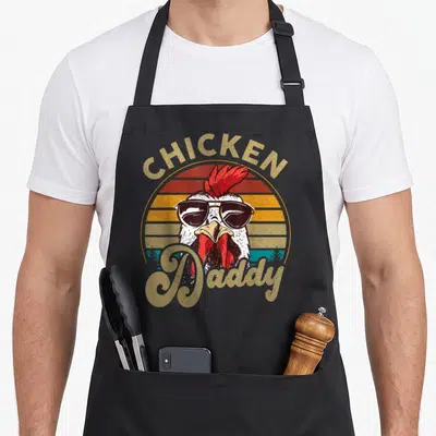 Farmer rooster apron - Retro chicken wearing sunglasses with CHICKEN Daddy text and striped sun background — shown on a black cotton canvas apron against a white background