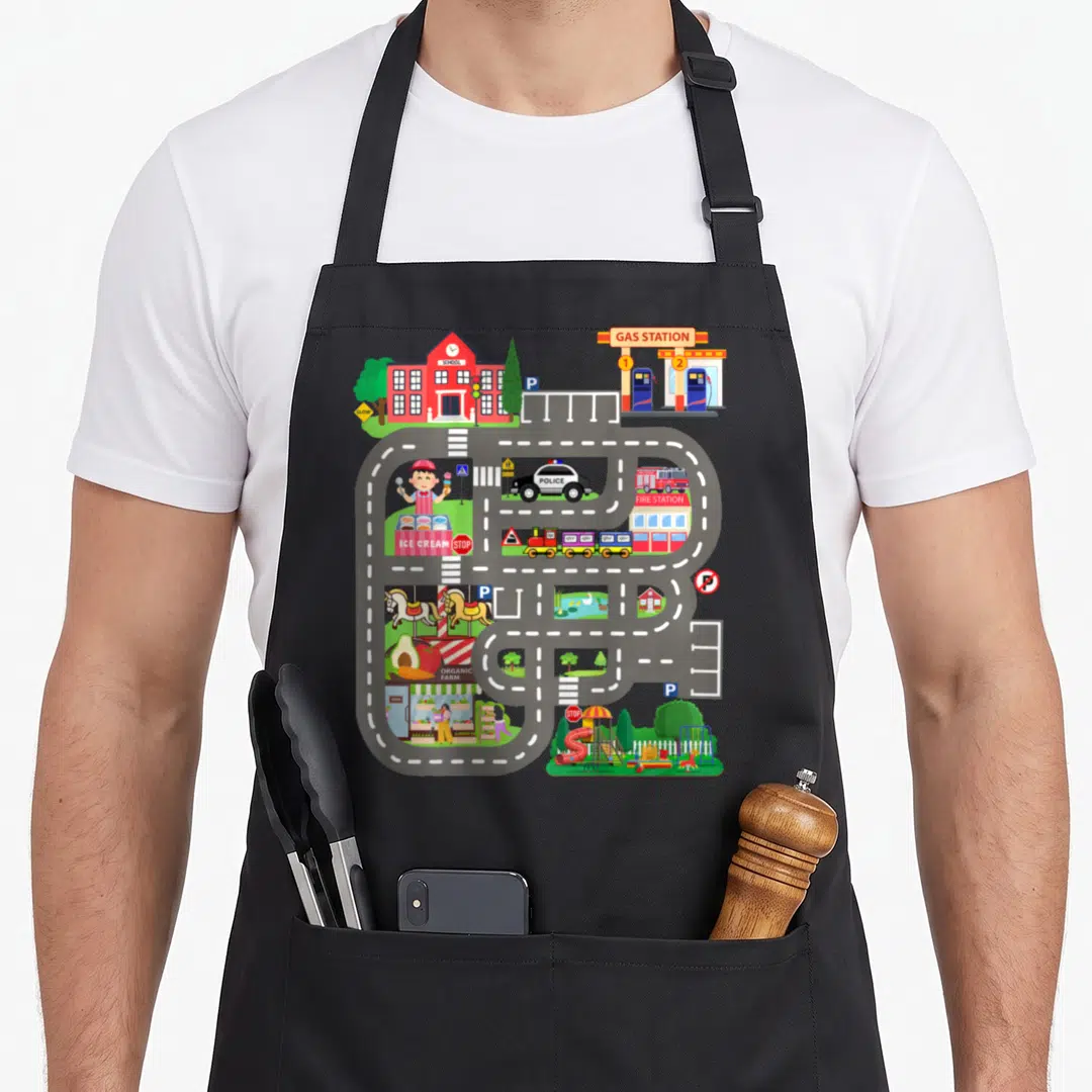 Father track apron - Toy town map with winding grey road connecting labeled buildings: school, gas station, police — shown on a black cotton canvas apron against a white background