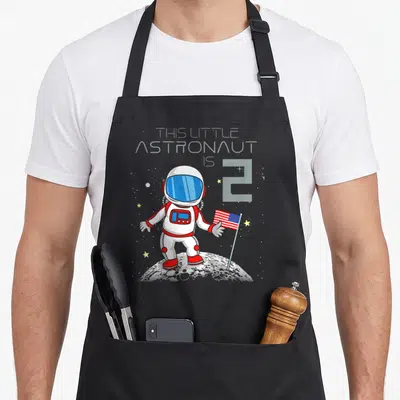 2nd geek apron - Cartoon astronaut holding a flag on the moon with text This Little Astronaut Is 2 — shown on a black cotton canvas apron against a white background