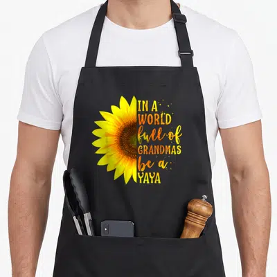Grandmas yaya mother's day aprons - Half sunflower slice next to stacked yellow and orange text reading IN A WORLD full — shown on a black cotton canvas apron against a white background