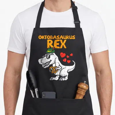 Oktobasaurus oktoberfest apron - Cartoon T-Rex in green hat holding orange pretzel with red hearts and stacked orange — shown on a black cotton canvas apron against a white background