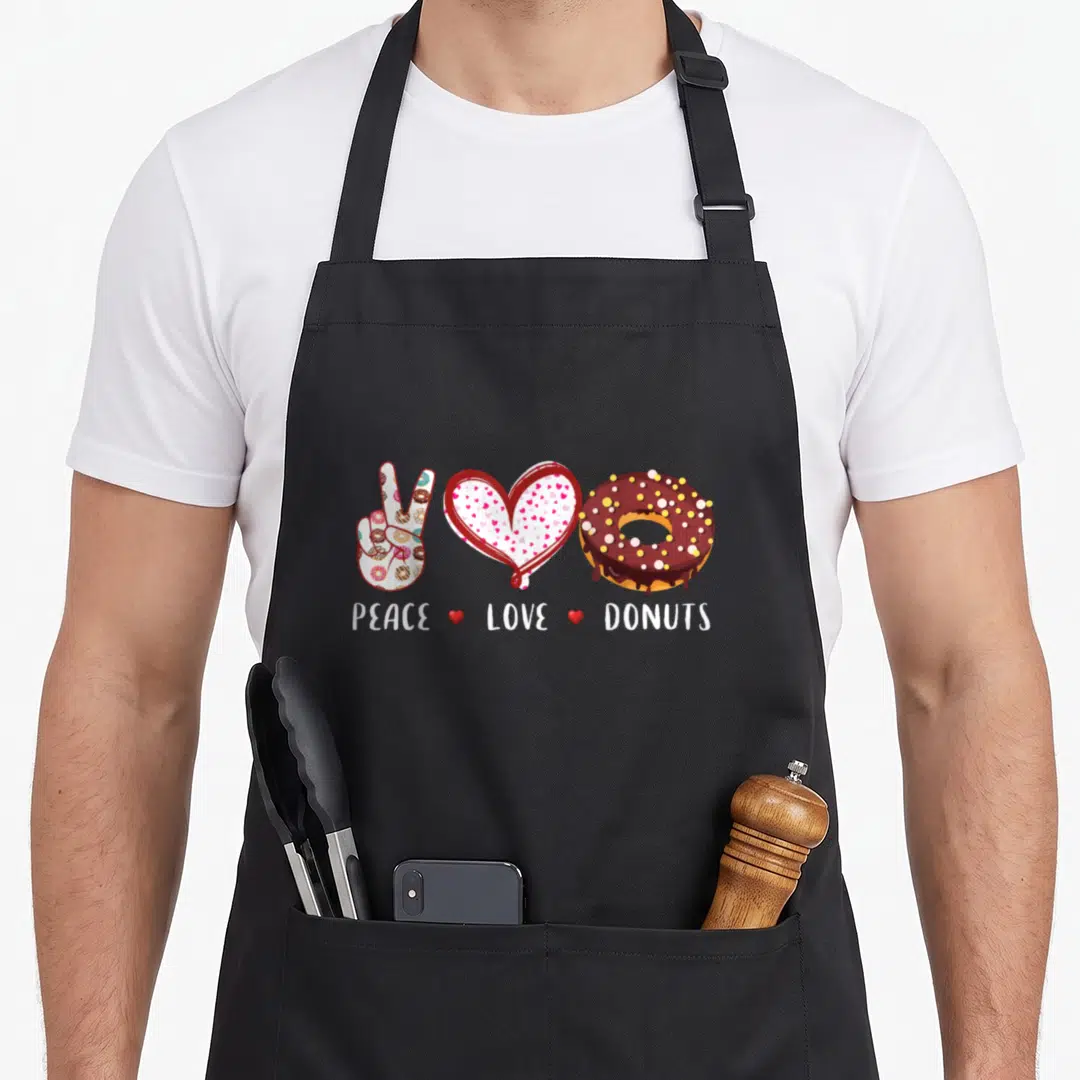 Peace donut apron - Cartoon peace sign, patterned heart, and chocolate donut with white text below — shown on a black cotton canvas apron against a white background