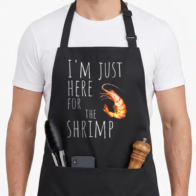 Shrimp i love food apron - Stacked white text I'M JUST HERE FOR THE SHRIMP beside an orange watercolor shrimp — shown on a black cotton canvas apron against a white background