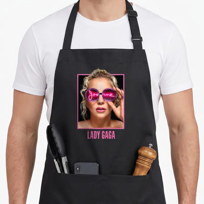 Gaga novelty apron - Woman wearing sunglasses with Joanne script inside lenses and LADY GAGA text below in pink frame — shown on a black cotton canvas apron against a white background