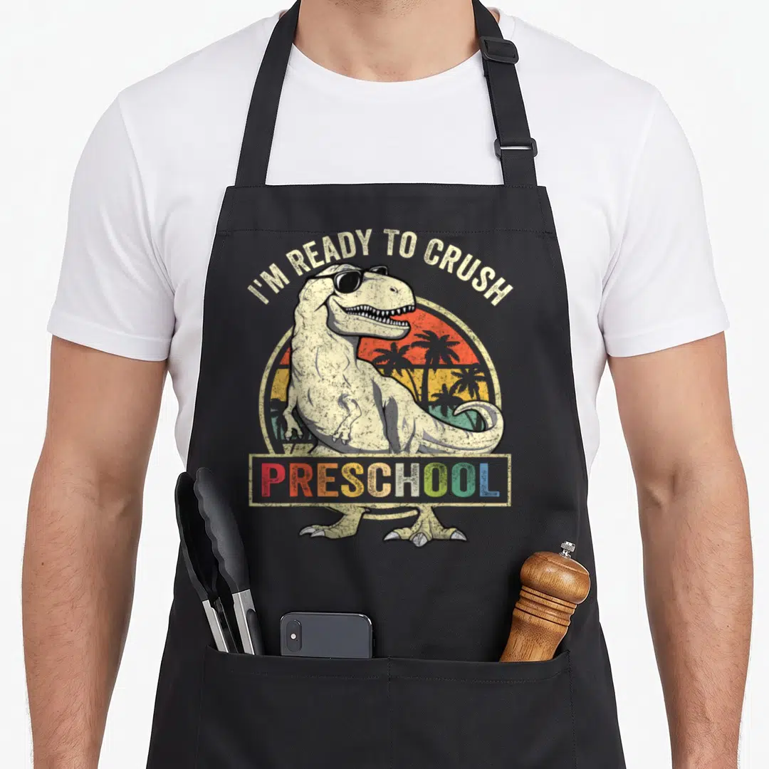 Crush preschool dinosaur apron - Retro Tyrannosaurus Rex in sunglasses with I'm Ready to Crush Preschool text — shown on a black cotton canvas apron against a white background