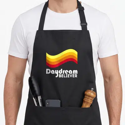 Daydream lettering quotes apron - Stacked Daydream BELIEVER text below a wavy multi-colored graphic ribbon — shown on a black cotton canvas apron against a white background