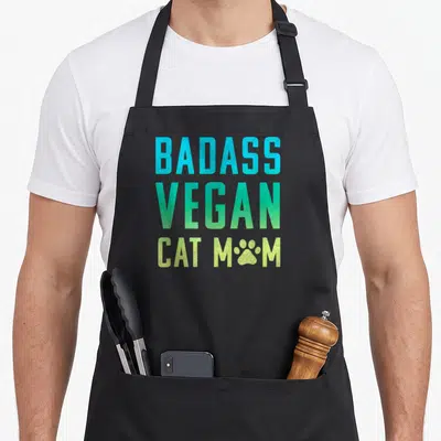 Vegan badass apron - Stacked BADASS VEGAN CAT MOM text in cyan green and lime with paw print replacing O — shown on a black cotton canvas apron against a white background