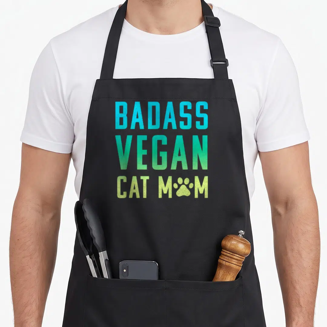 Vegan badass apron - Stacked BADASS VEGAN CAT MOM text in cyan green and lime with paw print replacing O — shown on a black cotton canvas apron against a white background