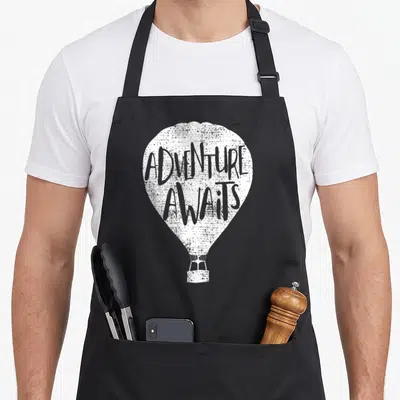 Aviator balloon apron - Distressed white hot air balloon with ADVENTURE AWAITS text on envelope — shown on a black cotton canvas apron against a white background