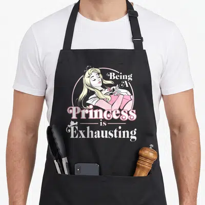 Sleeping princess apron - Cartoon blonde princess in pink circle with text Being A Princess is Exhausting — shown on a black cotton canvas apron against a white background