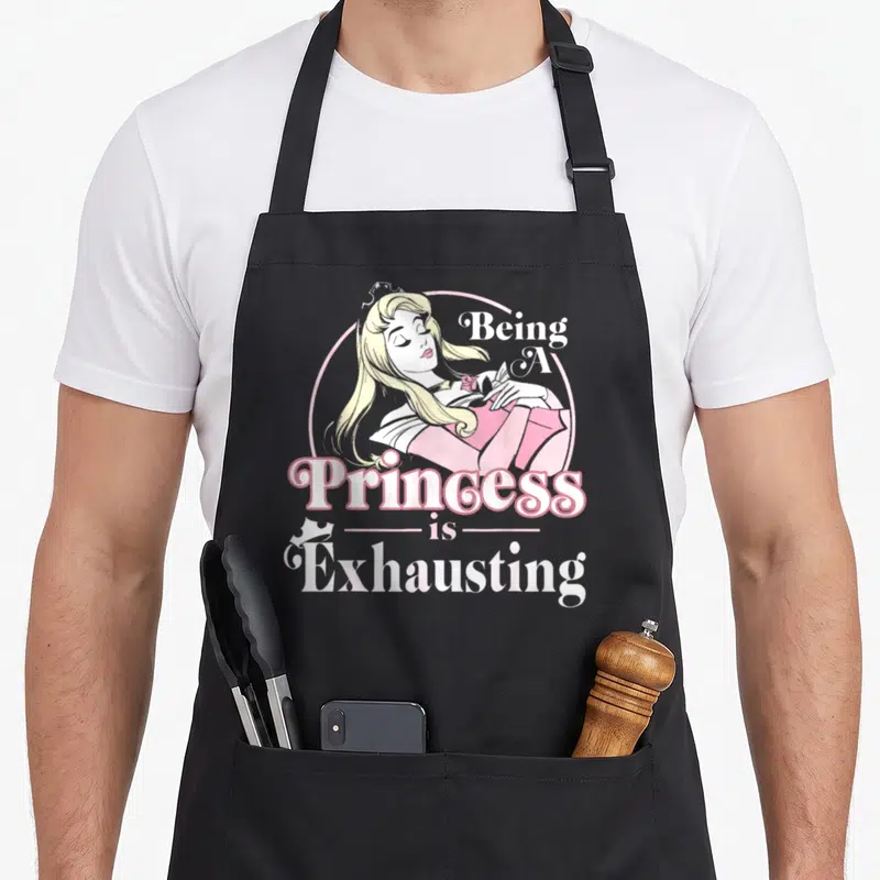 Sleeping princess apron - Cartoon blonde princess in pink circle with text Being A Princess is Exhausting — shown on a black cotton canvas apron against a white background