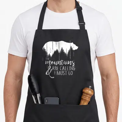 Calling bear apron - White bear silhouette containing grey pine trees with stacked inspirational text below — shown on a black cotton canvas apron against a white background