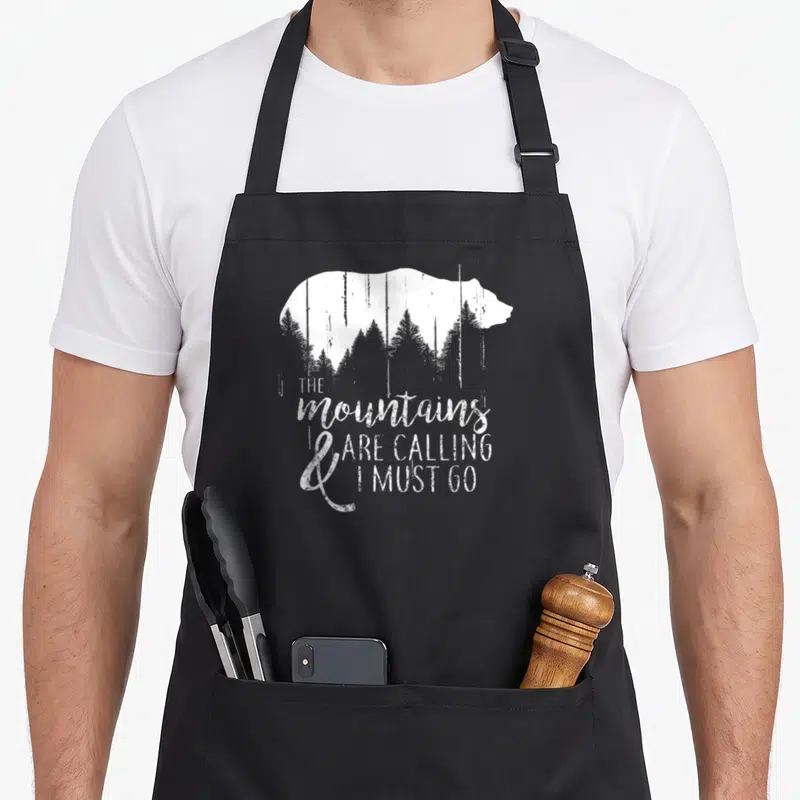 Calling bear apron - White bear silhouette containing grey pine trees with stacked inspirational text below — shown on a black cotton canvas apron against a white background