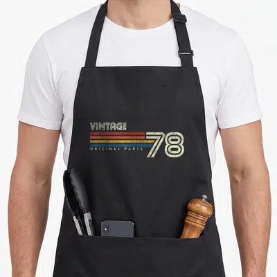 45th crew birthday apron - VINTAGE 78 ORIGINAL PARTS text with four colored stripes in distressed style — shown on a black cotton canvas apron against a white background
