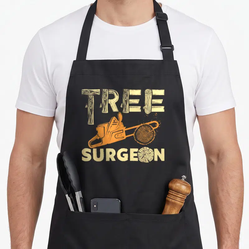 Tree arborist surgeon apron - Orange chainsaw cutting wood slice between tan log-style words TREE SURGEON — shown on a black cotton canvas apron against a white background