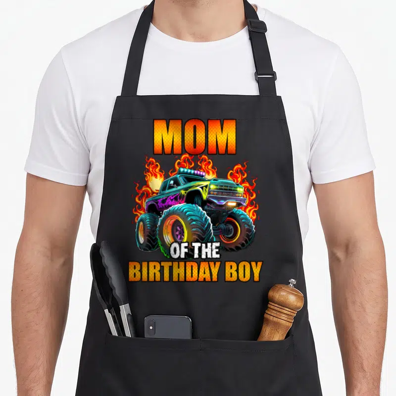 Cool birthday monster truck apron - MOM OF THE BIRTHDAY BOY text with cartoon monster truck and orange flames — shown on a black cotton canvas apron against a white background