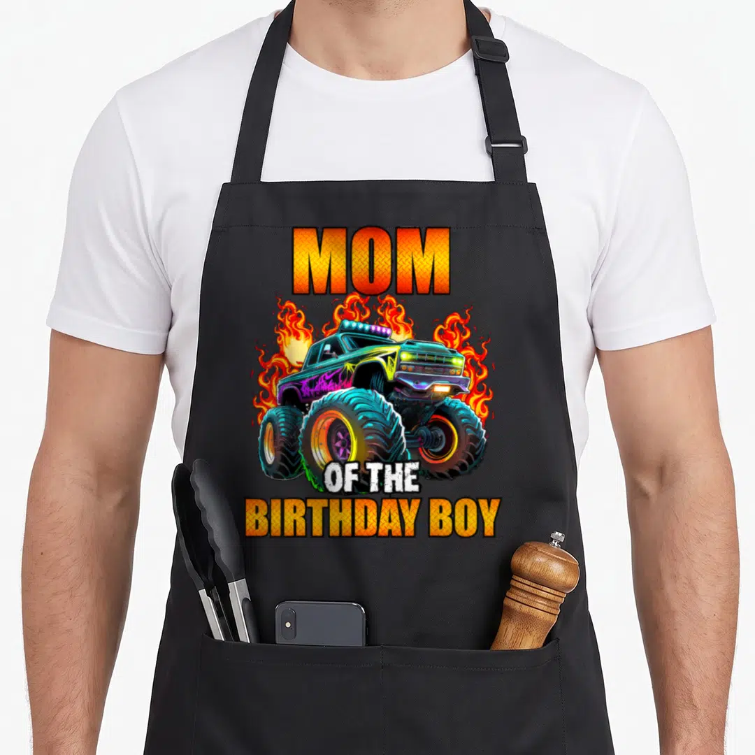 Cool birthday monster truck apron - MOM OF THE BIRTHDAY BOY text with cartoon monster truck and orange flames — shown on a black cotton canvas apron against a white background