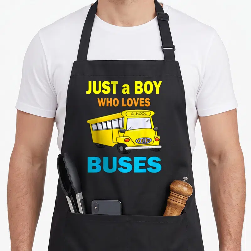 Buse school bus apron - Cartoon yellow school bus with text JUST a BOY WHO LOVES SCHOOL BUSES in yellow orange and blue — shown on a black cotton canvas apron against a white background