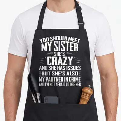 Sister nun apron - White stacked typography reading You Should Meet My Sister She's Crazy Partner In Crime — shown on a black cotton canvas apron against a white background
