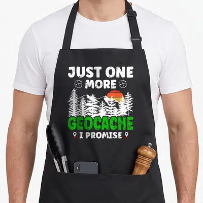 Geocache geocaching apron - Just One More Geocache I Promise text with white forest silhouette, orange sun, and green — shown on a black cotton canvas apron against a white background