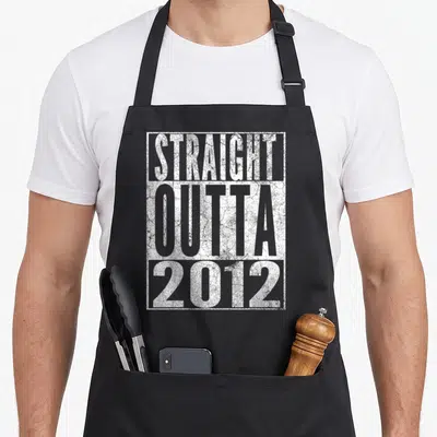 Outta 14th birthday apron - White distressed text STRAIGHT OUTTA 2012 stacked in rectangle on black background — shown on a black cotton canvas apron against a white background