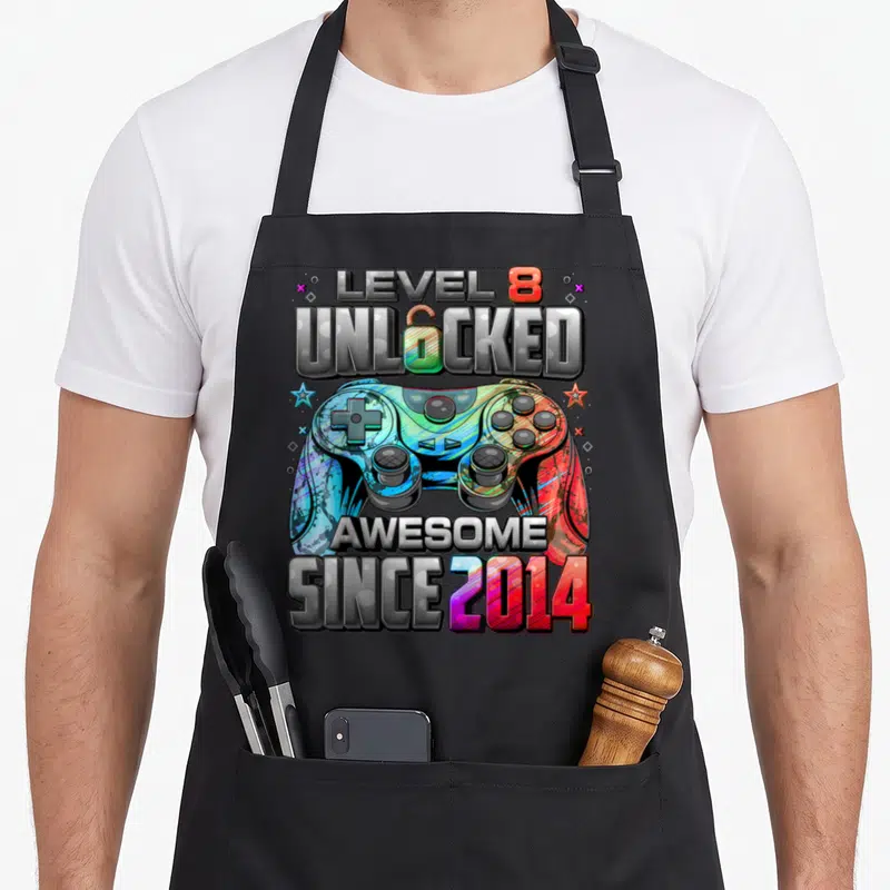 8th birthday gaming aprons - Colorful game controller with Level 8 Unlocked text and stars flanking device — shown on a black cotton canvas apron against a white background