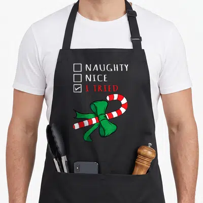Funny christmas candy cane apron - Cartoon checklist with NAUGHTY NICE I TRIED checked and red white candy cane — shown on a black cotton canvas apron against a white background