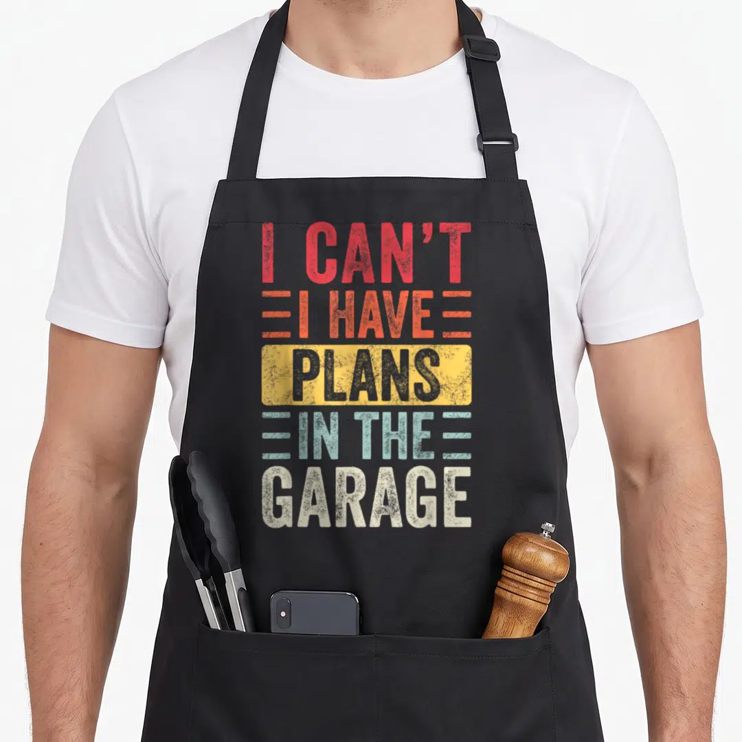 Cute plan garage apron - Stacked text reading I CAN'T I HAVE PLANS IN THE GARAGE in red orange yellow and blue — shown on a black cotton canvas apron against a white background