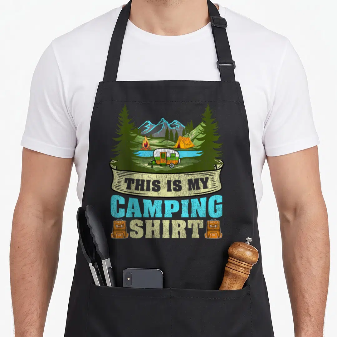 Camper travel trailer apron - Cartoon camping scene with trailer, tent, fire, mountains, and text THIS IS MY CAMPING — shown on a black cotton canvas apron against a white background