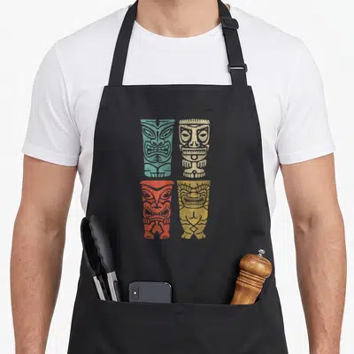 Polynesian tiki aprons - Four distressed tiki poles in teal, beige, red, and yellow arranged in a grid — shown on a black cotton canvas apron against a white background